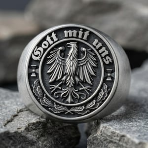 May include: A silver-toned metal ring featuring a circular face with an embossed eagle at the center. The text "Gott mit uns" is engraved along the upper rim, flanked by two small crosses, with oak leaf detailing at the base of the eagle.