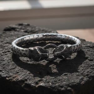 May include: A silver bracelet with a panther head design. The bracelet features two panther heads encrusted with black gemstones that connect with a silver clasp. The band has a honeycomb pattern. The bracelet is displayed on a dark, textured surface.