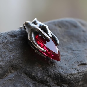 May include: A silver-toned pendant necklace featuring a large, faceted, red gemstone held by two detailed hands. The hands appear to be made of metal and are intricately sculpted. The gemstone is a deep, rich red color.