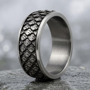 Puede incluir: Un anillo ancho de metal con un acabado oscuro y oxidado que presenta un patrón repetitivo en relieve similar a escamas en la banda exterior. El interior del anillo tiene un acabado plateado cepillado y liso, que contrasta con el diseño texturizado.
