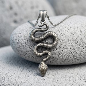 以下が含まれることがあります： 銀色の金属製ヘビのペンダントで、お揃いの細いリンクチェーンから吊り下げられています。ヘビはS字型に巻いた状態で表現されており、体全体が詳細な質感のある鱗で覆われ、上部には尖ったバチカンが付いています。