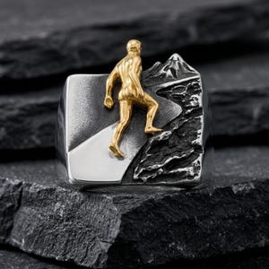 Puede incluir: Un anillo plateado con una figura dorada de una persona escalando una montaña. El anillo tiene una cara rectangular con un diseño de montaña en un lado y un camino en el otro. La figura está escalando la montaña.
