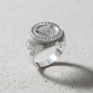 Puede incluir: Un anillo plateado con un diseño geométrico. El anillo presenta una cara circular con un triángulo en relieve y un borde de pequeñas piedras brillantes. La banda tiene un patrón texturizado en los lados. El anillo está sobre un fondo claro.