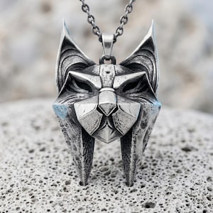 May include: A silver-toned metal pendant shaped like a stylized lynx or cat head with sharp, angular features and a textured, weathered finish. The pendant hangs from a matching silver-toned metal chain, showcasing intricate geometric detailing.