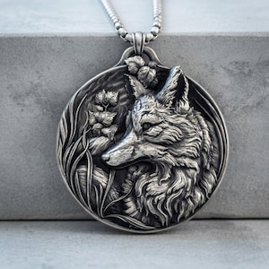 May include: A circular silver pendant featuring a detailed relief of a fox head surrounded by foliage and tall grass. The pendant has an oxidized finish to highlight the texture of the fur and leaves, and it hangs from a silver chain necklace.
