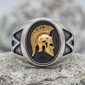 Puede incluir: Un anillo de tono plateado con una incrustación ovalada central sobre un fondo oscuro y texturizado. Un casco espartano de color dorado pulido con una cresta prominente se centra en la incrustación. El anillo tiene un acabado cepillado.