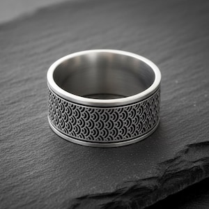 May include: A wide, silver-colored ring with a detailed, repeating wave pattern around the band. The ring sits on a dark, textured surface, showcasing its intricate design and metallic sheen. A modern piece of jewelry.