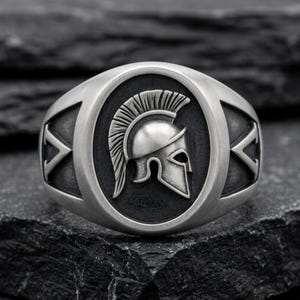 Puede incluir: Anillo plateado con un diseño detallado de casco espartano dentro de un marco ovalado. El anillo tiene una banda ancha con detalles decorativos a cada lado. El casco tiene una cresta prominente y está sobre un fondo oscuro.