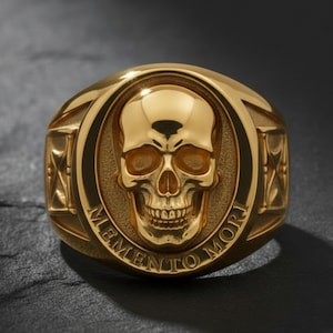 May include: A gold-colored ring featuring a detailed skull design in the center. The ring has the words "MEMENTO MORI" engraved around the skull. The ring has a polished finish and is set against a dark background.