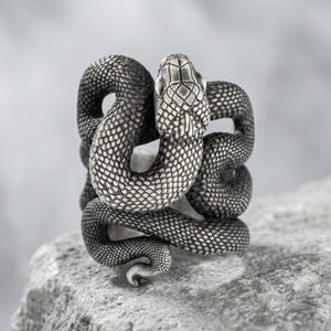 Op de afbeelding: Een zilverkleurige ring in de vorm van een opgerolde slang met gedetailleerde schubben over het hele lichaam. De kop van de slang bevindt zich aan de bovenkant met een textuurpatroon, terwijl het lichaam de ringband vormt.
