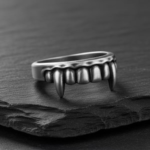 May include: A silver-colored ring featuring a row of teeth with two prominent fangs. The ring is set against a dark, textured surface, creating a striking contrast. The design evokes a gothic or Halloween theme.