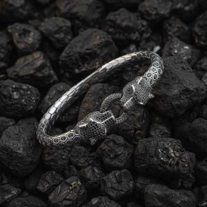 Könnte beinhalten: Ein silbernes Armband mit zwei Pantherköpfen, die mit schwarzen Edelsteinen besetzt sind. Das Armband hat ein strukturiertes, sechseckiges Muster entlang des Bandes. Die Pantherköpfe verbinden sich zu einem Verschluss, wodurch ein einzigartiges Schmuckstück entsteht.