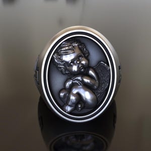 May include: A silver ring with a detailed cherub sculpture in the center. The cherub is sitting with its wings folded behind it.