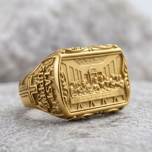 May include: A gold-toned ring featuring a rectangular face with a relief carving of The Last Supper. The side of the band displays a cross and the text LAST SUPPER in raised lettering. The metal surface has a polished finish with intricate details.