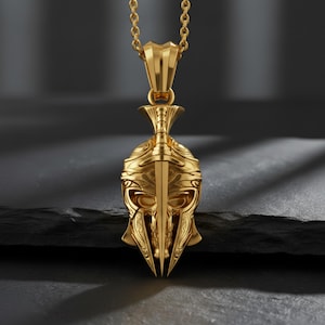 May include: A gold-colored pendant necklace featuring a detailed, stylized helmet design. The pendant has intricate carvings and is suspended from a matching gold chain. The helmet design includes a central sword element.