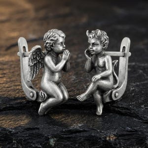 May include: A pair of silver-toned earrings featuring angel and devil figures. The angel is kneeling with hands clasped in prayer, while the devil sits with a hand to its face. The earrings have a unique, detailed design.