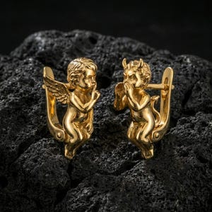 Puede incluir: Un par de pendientes dorados. Uno de los pendientes presenta un querubín con alas, el otro un diablo con cuernos. Ambas figuras están sentadas con un dedo en los labios, en un gesto de silencio. Los pendientes están sobre un fondo oscuro y texturizado.