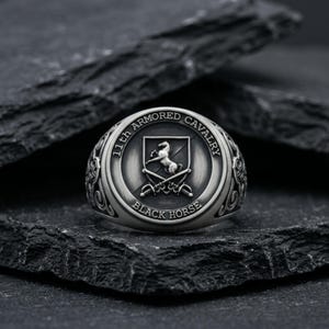 May include: Silver-toned ring with a detailed design. The ring features the text "11th ARMORED CAVALRY BLACK HORSE" around a central emblem of a horse and crossed swords. The ring is set against a dark, textured background.