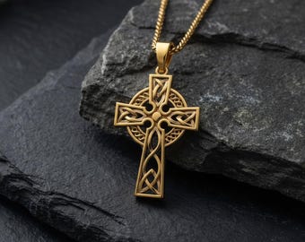 Op de afbeelding: Een goudkleurige Keltische kruis hanger met ingewikkeld knoopwerk, hangend aan een bijpassende ketting. De hanger wordt getoond tegen een donkere, getextureerde stenen achtergrond, die het gedetailleerde vakmanschap benadrukt.