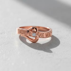 May include: Rose gold ring with the inscription "TRUE LOVE WAITS". The ring features a heart-shaped design with a small, clear gemstone in the center. The ring is on a white surface.