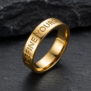 Puede incluir: Un anillo dorado con las palabras grabadas "DEFINE YOURSELF". El anillo está hecho de un metal brillante y reflectante y está sobre un fondo oscuro y texturizado. La inscripción del anillo es claramente visible.