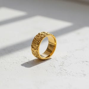 以下が含まれることがあります： 金色のリングで、表面はテクスチャ加工されています。リングは、詳細で不規則なパターンを持つバンドのようなデザインです。リングは白い表面に表示され、影が深さを加えています。