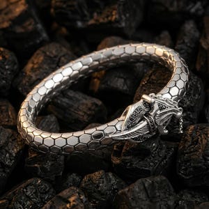 Könnte beinhalten: Ein silbernes Armband mit einem Drachenkopfverschluss. Das Armband hat ein sechseckiges Muster und ist vor einem dunklen Hintergrund platziert. Der Drachenkopf ist mit schwarzen Akzenten versehen.