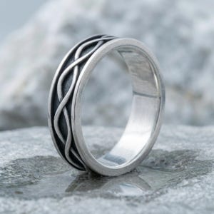 May include: A silver-toned metal ring featuring a continuous, raised, intertwined cable or rope pattern around the outer band. The recessed areas between the raised silver lines are darkened, creating a high-contrast effect against the polished edges.