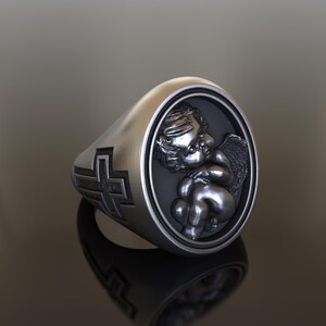 May include: A silver ring with a cross on one side and a cherub with wings on the other side.