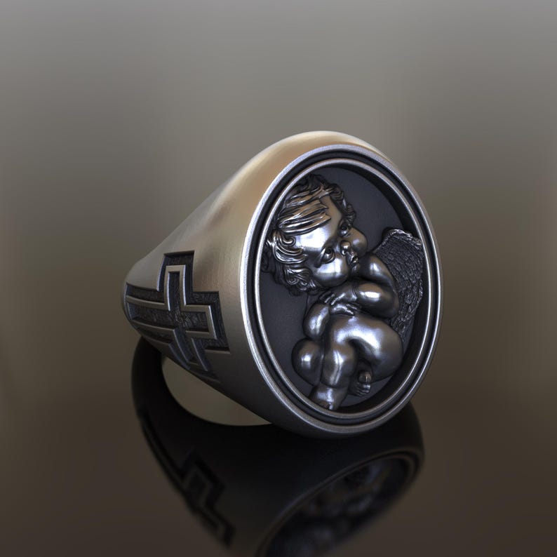 May include: A silver ring with a cross on one side and a cherub with wings on the other side.