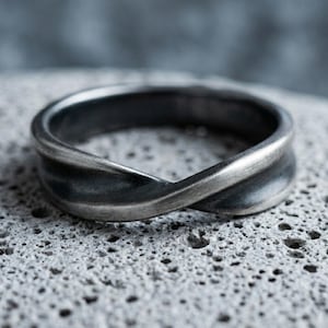 Peut inclure: Une bague en métal gris foncé avec un design torsadé rappelant le symbole de l'infini. L'anneau présente une finition brossée et mate avec des zones plus sombres en creux qui soulignent les courbes du métal, créant une forme fluide continue.