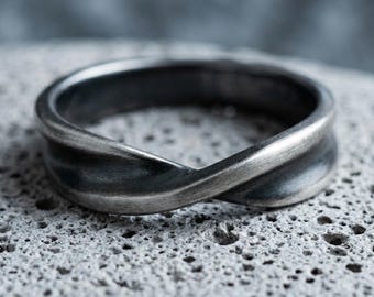 May include: A dark gray metal ring with a twisted, infinity-like design. The band features a brushed, matte finish with darker recessed areas that highlight the curves of the metal, creating a continuous, flowing shape around the entire circumference.