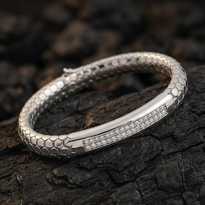 Peut inclure: Un bracelet jonc argenté avec un motif texturé en nid d'abeille. Une section du bracelet est lisse et sertie de deux rangées de petits pierres précieuses transparentes. Le bracelet est présenté sur une surface sombre et texturée.