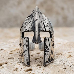Op de afbeelding: Een zilverkleurige metalen ring in de vorm van een Spartaanse helm. Het oppervlak heeft een verweerde afwerking met donkere, verdiepte delen en een puntige kam. Het ontwerp bevat een centrale neusbeschermer en twee zijdelingse wangplaten.