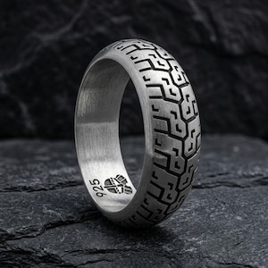 May include: A silver ring with a tire tread pattern. The ring is made of a brushed metal and features a repeating geometric design. The ring is marked with "925" and a small emblem.