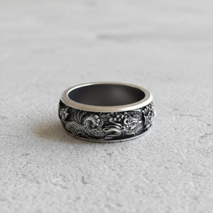 May include: A silver ring with a detailed dragon design. The ring has a dark interior and a wide band with an embossed dragon motif. The dragon is depicted with intricate scales and flowing details, creating a bold and artistic statement.
