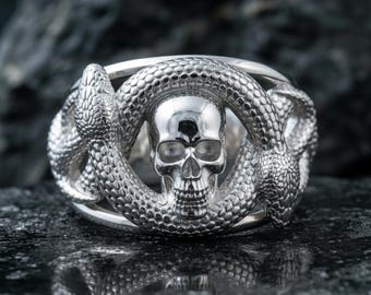 May include: A silver ring featuring a skull encircled by two snakes. The skull has detailed facial features, and the snakes have textured scales. The ring is set against a dark, textured background.