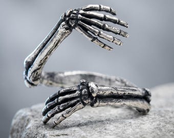 May include: A silver-toned metal ring featuring a wrap-around design shaped like two skeletal hands. The detailed finish highlights the texture of the finger bones and wrist joints, creating a continuous band that encircles the finger with an open gap.