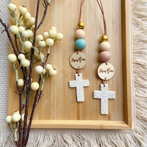 May include: Two wooden cross charms with a white textured design and a wooden bead necklace with light blue, pink and cream coloured beads. The charms have a wooden tag with the text "Jesus loves me" on them.