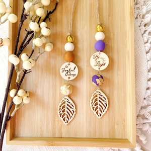 May include: Two decorative hanging ornaments with wooden beads, leaf cutouts, and inspirational quotes. One ornament says "be joyful always" and the other says "Be the Light". The beads are gold, white, brown, and purple.
