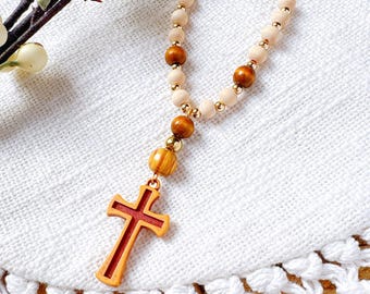 May include: A wooden rosary necklace with a gold chain and cross pendant. The necklace features light beige and brown wooden beads, with a red cross at the bottom. The rosary is displayed on a white, textured surface.