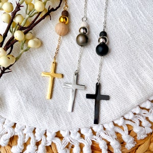 May include: Three silver and gold necklaces with cross pendants and wooden and metal beads. The necklaces are hanging on a white fabric background.