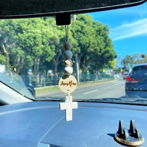 May include: A wooden car charm with a cross and a circle that says "Jesus ♥ Me" hanging from a rearview mirror. The charm is made of light brown wood and has a white and beige beaded chain.