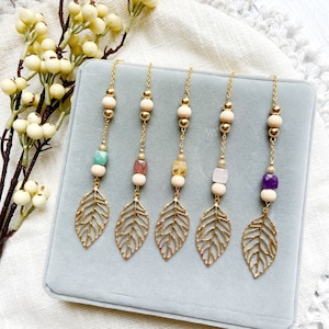 May include: Five gold chains with leaf-shaped charms and various coloured gemstone beads. The chains are displayed on a light blue velvet surface.