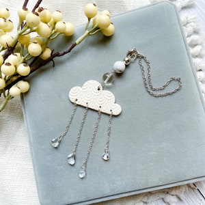 May include: White ceramic cloud charm with a silver chain and clear crystal raindrops hanging from it. The charm is on a grey velvet background.