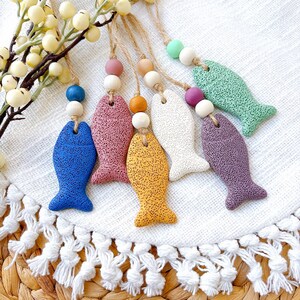 May include: A selection of vibrantly coloured, textured fish-shaped ornaments. Each fish is a different hue, including blue, pink, yellow, white, green, and purple. They are strung with wooden beads and twine, suggesting they are for hanging.
