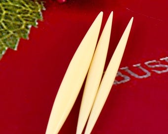 May include: Three ivory-colored, pointed tools are arranged on a deep red surface. The tools are slender and tapered, with a smooth, glossy finish. The background includes a hint of a green leaf and a silver-colored inscription.