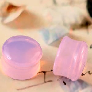 May include: Two pink and lavender ear plugs. One plug is round and the other is a curved, rectangular shape. The plugs are made of a translucent material. The background is a light tan colour with some blurred text.