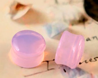 May include: Two pink and lavender ear plugs. One plug is round and the other is a curved, rectangular shape. The plugs are made of a translucent material. The background is a light tan color with some blurred text.