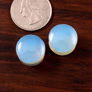 May include: Two round, light blue opal cabochons, each approximately 1 inch in diameter, on a brown wooden surface.